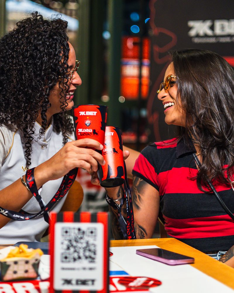 Duas amigas brindando com copos da 7K Bet à mesa do bar