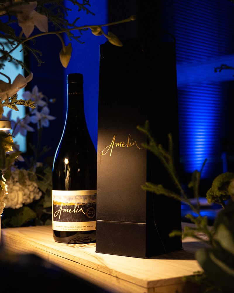 Garrafa de vinho Amelia ao lado da sacola da marca, sob luz azul